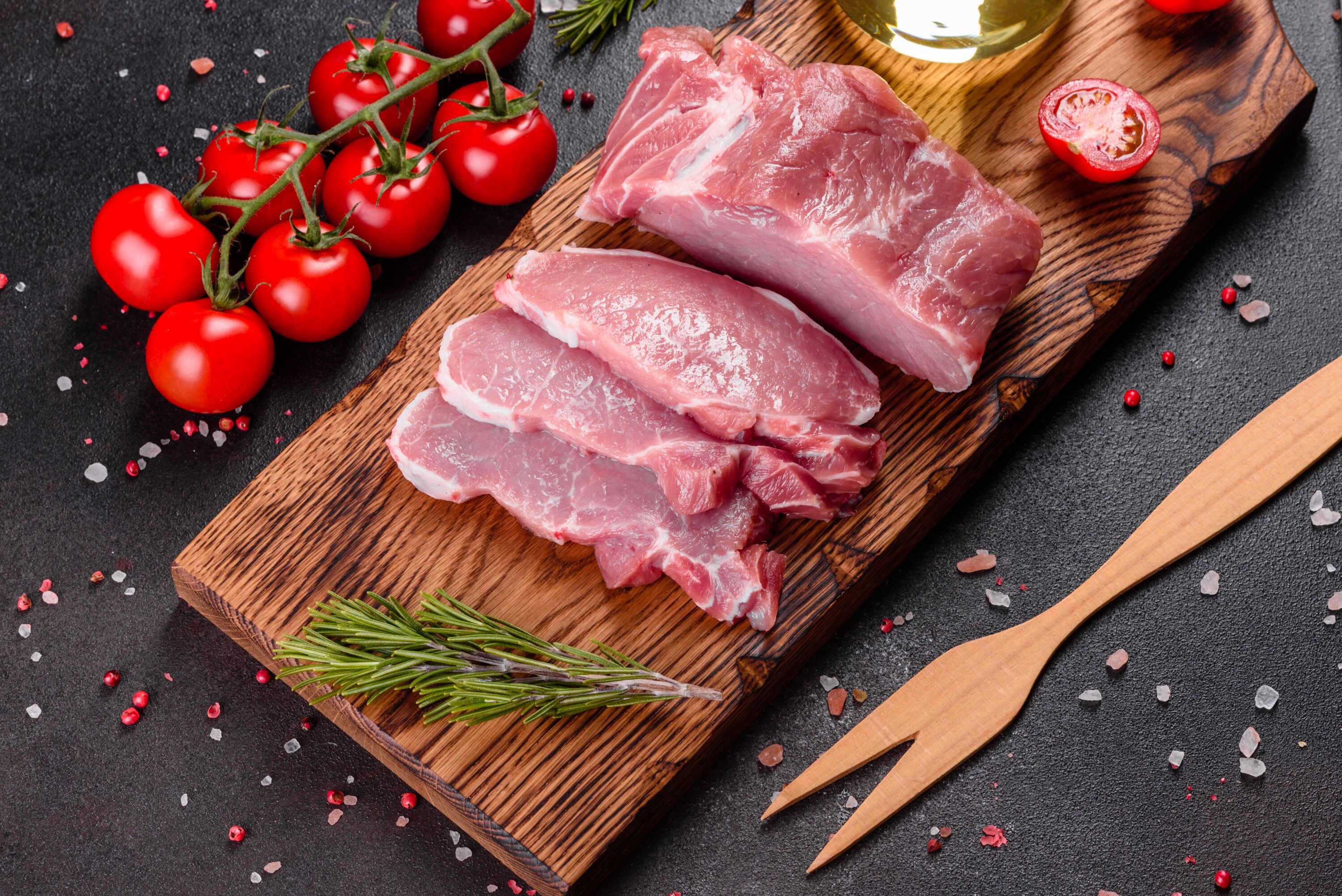 Свежесрубленное мясо на деревянной разделочной доске, украшенной розмарином, с рядом красными помидорами черри