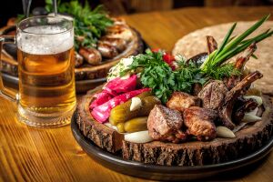 Аппетитное мясное ассорти на деревянной тарелке с хрустящими соленьями и свежей зеленью, сопровождаемое прохладным пивом
