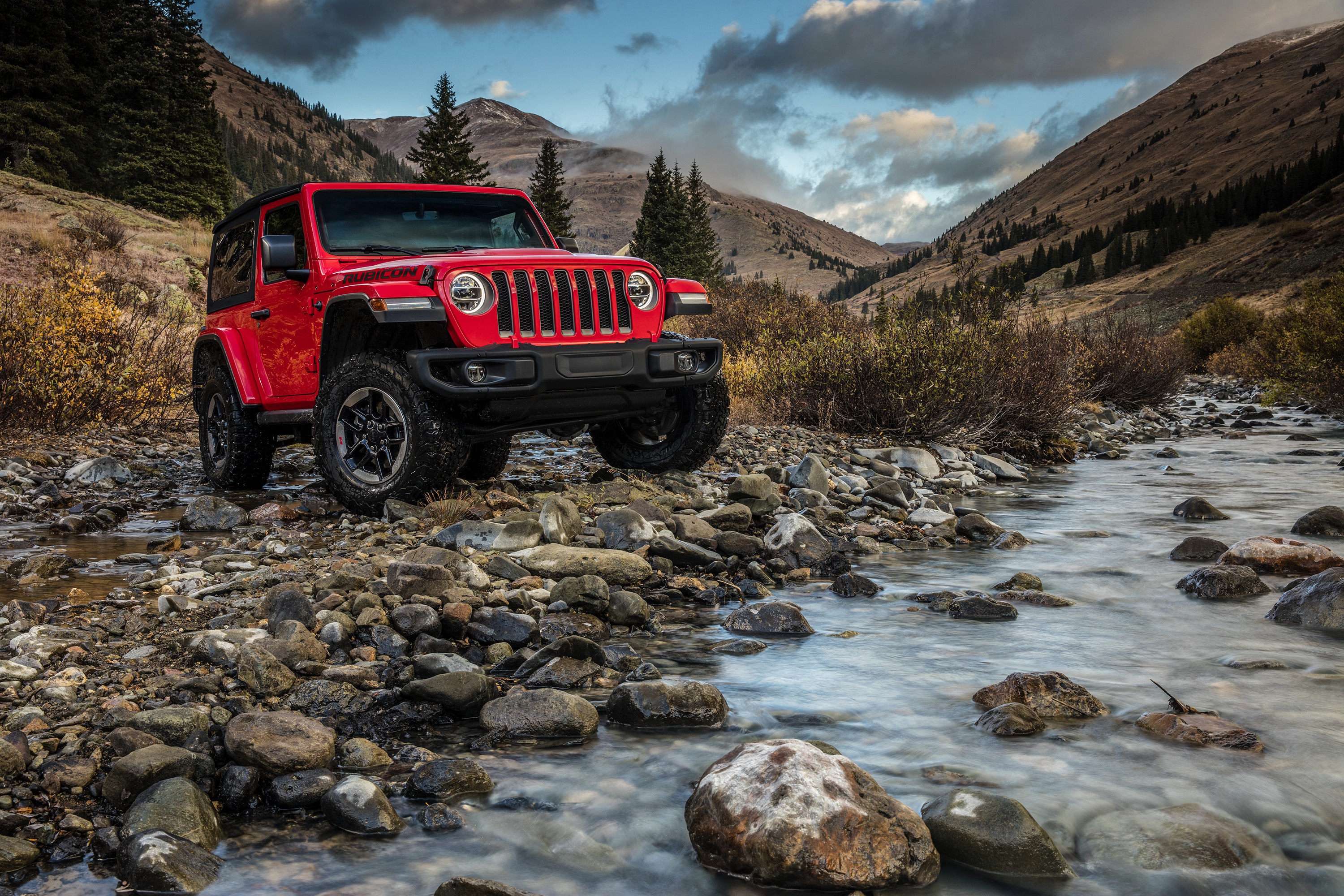 Красный Jeep Wrangler Rubicon, преодолевающий горную реку среди скалистых склонов и темного леса